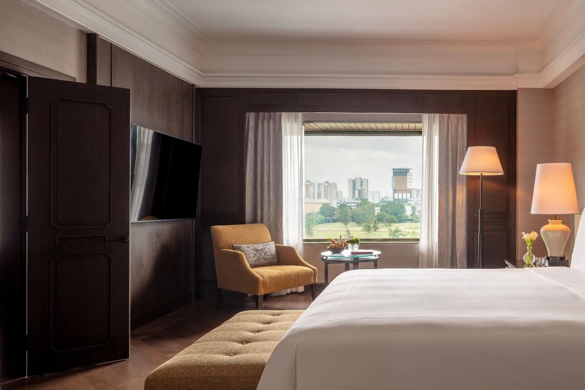 A hotel room with a large bed, a TV, a chair, a small table with a lamp, and a view of the city.