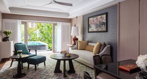 Habitación de hotel moderna con sofá, sillones, una mesa de centro, una mesa de comedor y vistas a una piscina y zonas verdes.