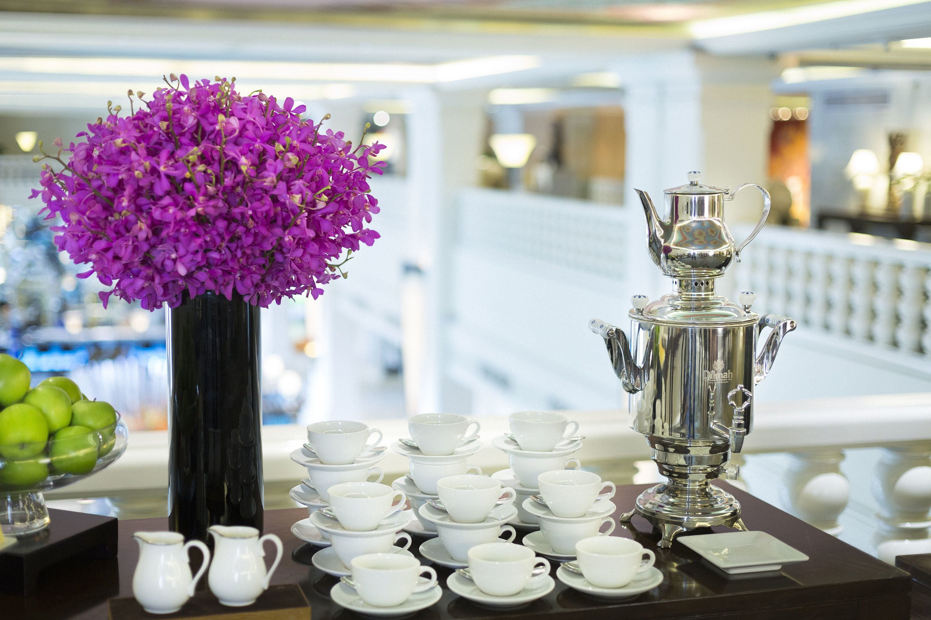Une table avec une théière en argent, plusieurs tasses et soucoupes blanches, et un vase rempli de fleurs violettes.