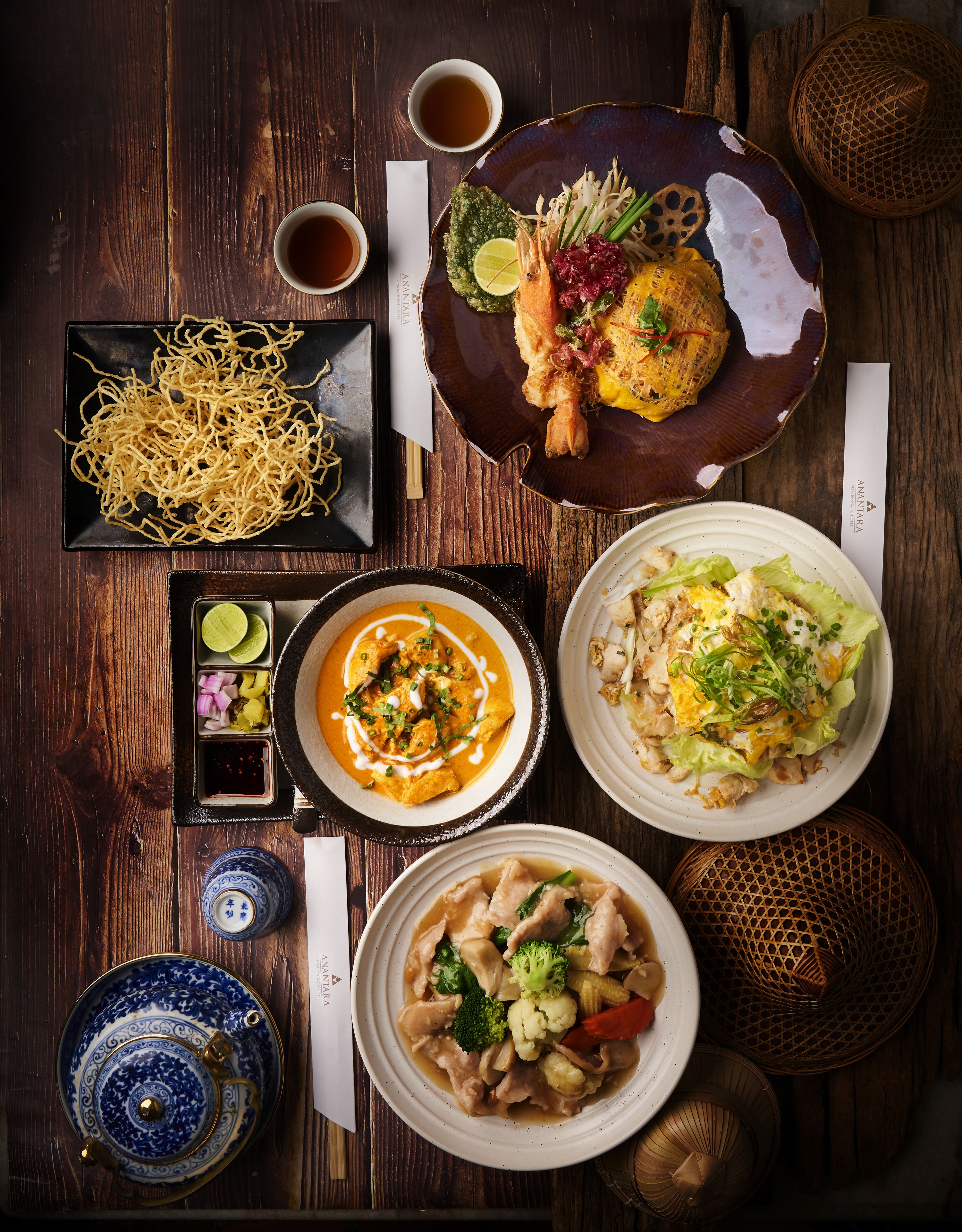 A variety of dishes on a wooden table, including noodles, soup, and a plate with a whole shrimp.