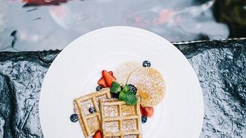 Une assiette de gaufres garnies de fraises et de myrtilles, posée sur une surface en pierre, près d’une étendue d’eau où l’on peut voir des poissons.