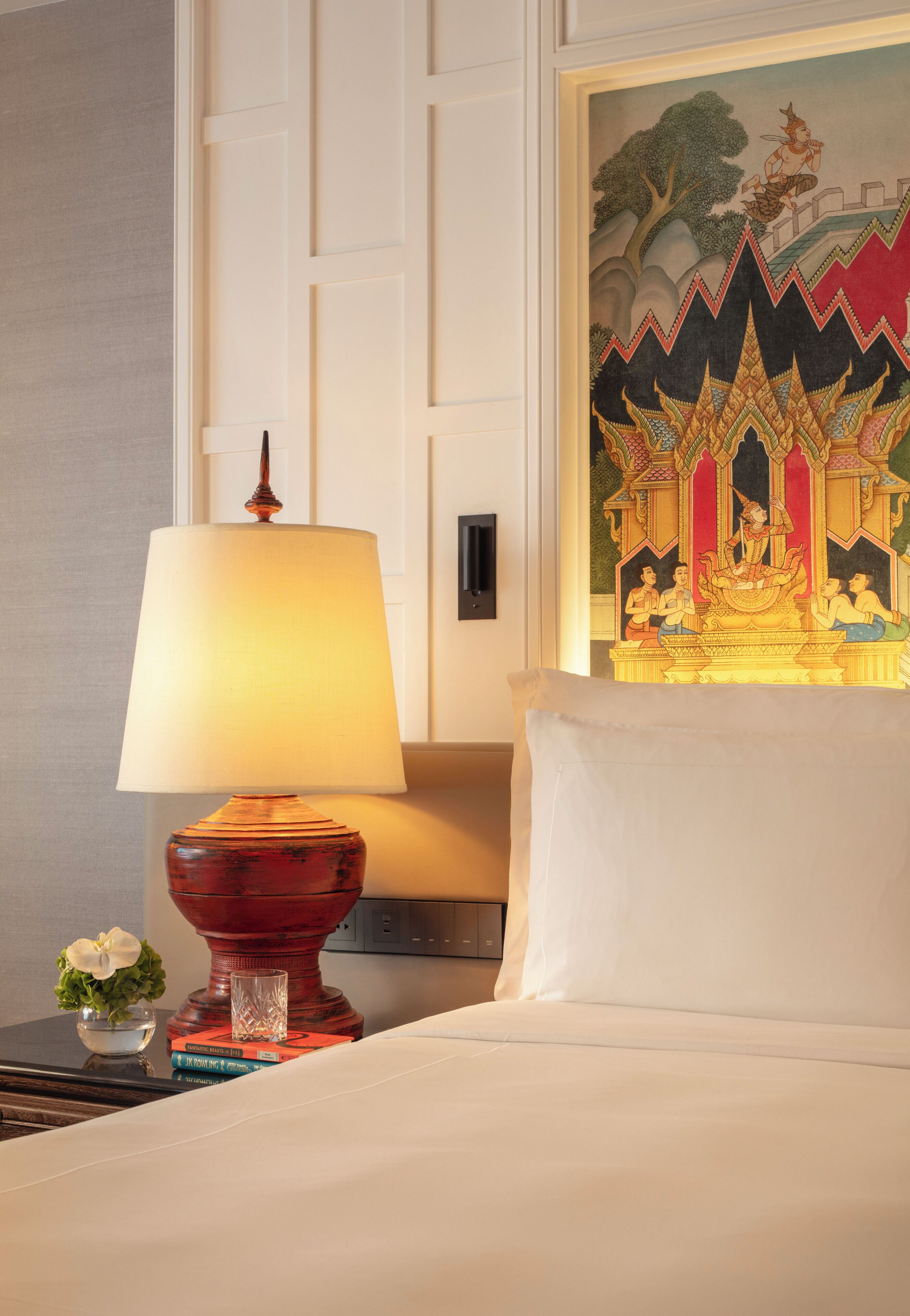 A hotel room with a bed, a lamp, a framed picture, and a small vase with a flower.