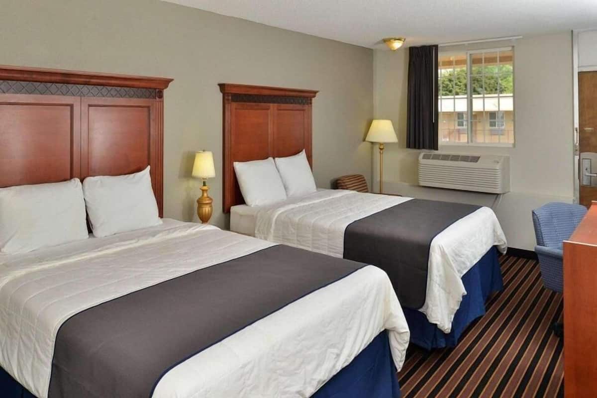 Quarto de hotel com duas camas, uma escrivaninha, uma cadeira, uma luminária, ar condicionado e uma janela com vista.