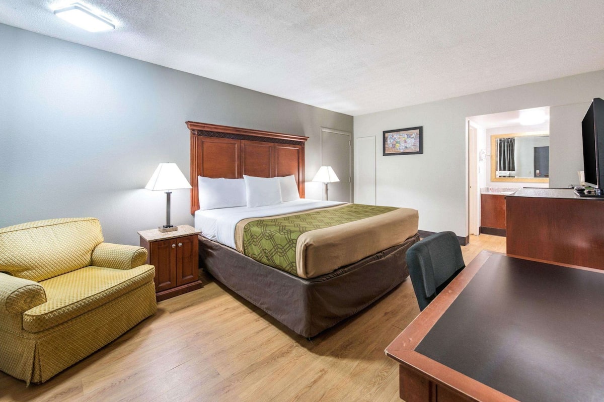 Quarto de hotel com cama, poltrona, escrivaninha e televisão.