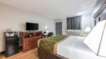 A hotel room with a bed, a desk with a computer, a television, a refrigerator, and an air conditioning unit.