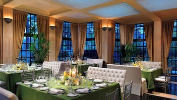 Une salle à manger élégante, avec plusieurs tables dressées pour un repas, de grandes fenêtres et un lustre.