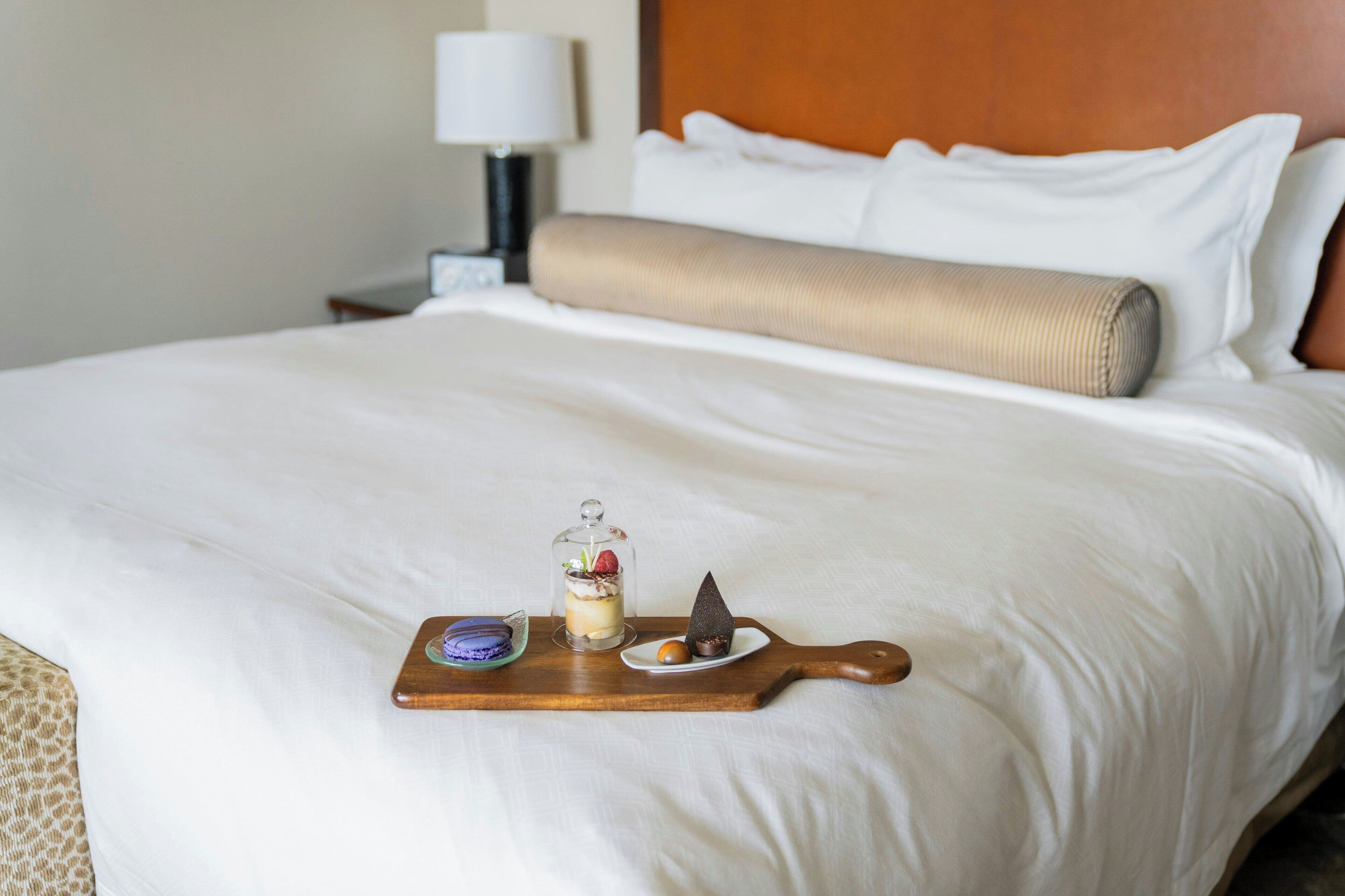 A neatly made bed with a wooden tray holding a dessert and a chocolate piece, a lamp, and a nightstand with a lamp.