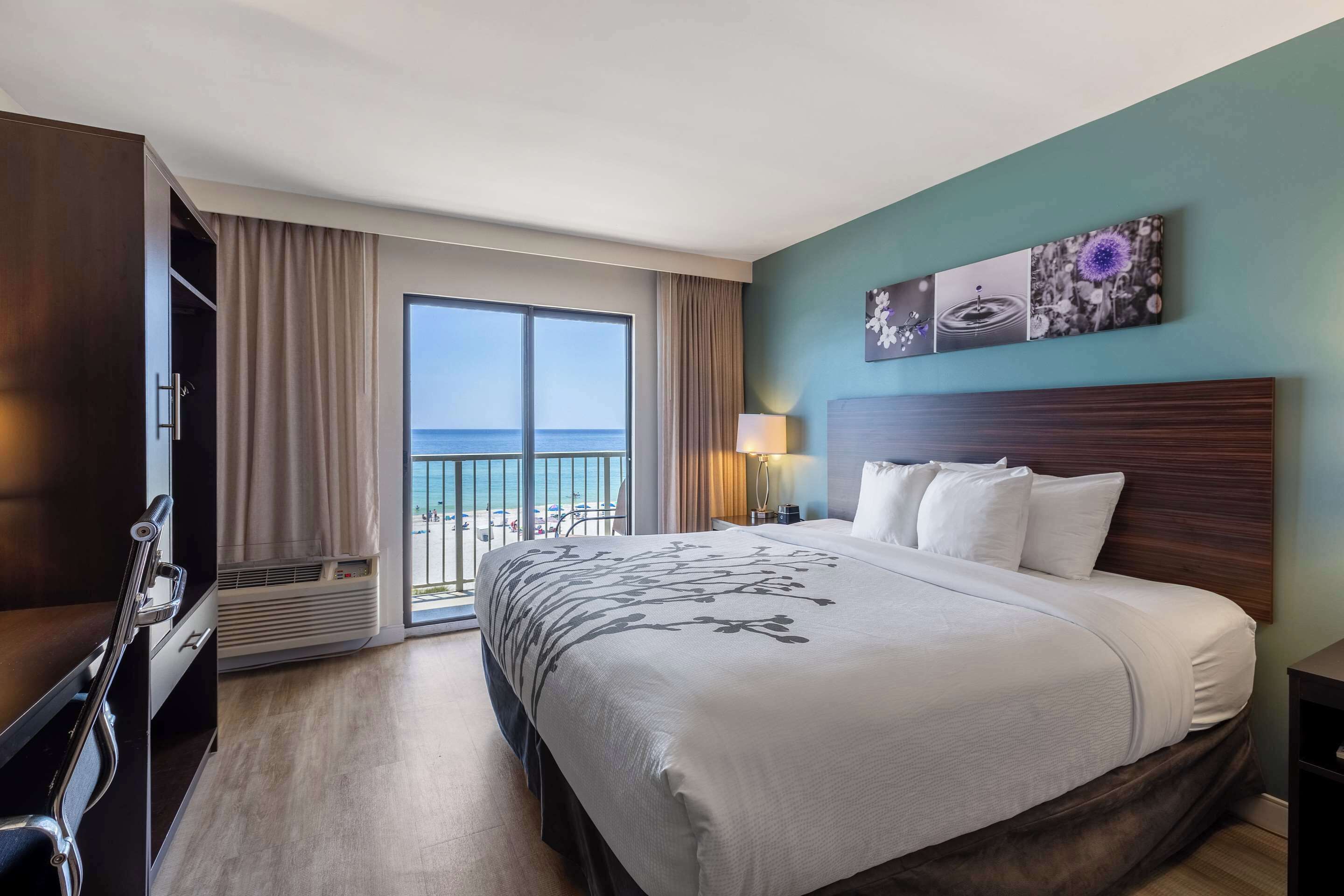 A hotel room with a large bed, a desk, a chair, a nightstand, a lamp, and a view of the ocean.