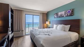 A hotel room with a large bed, a desk, a chair, a nightstand, a lamp, and a view of the ocean.