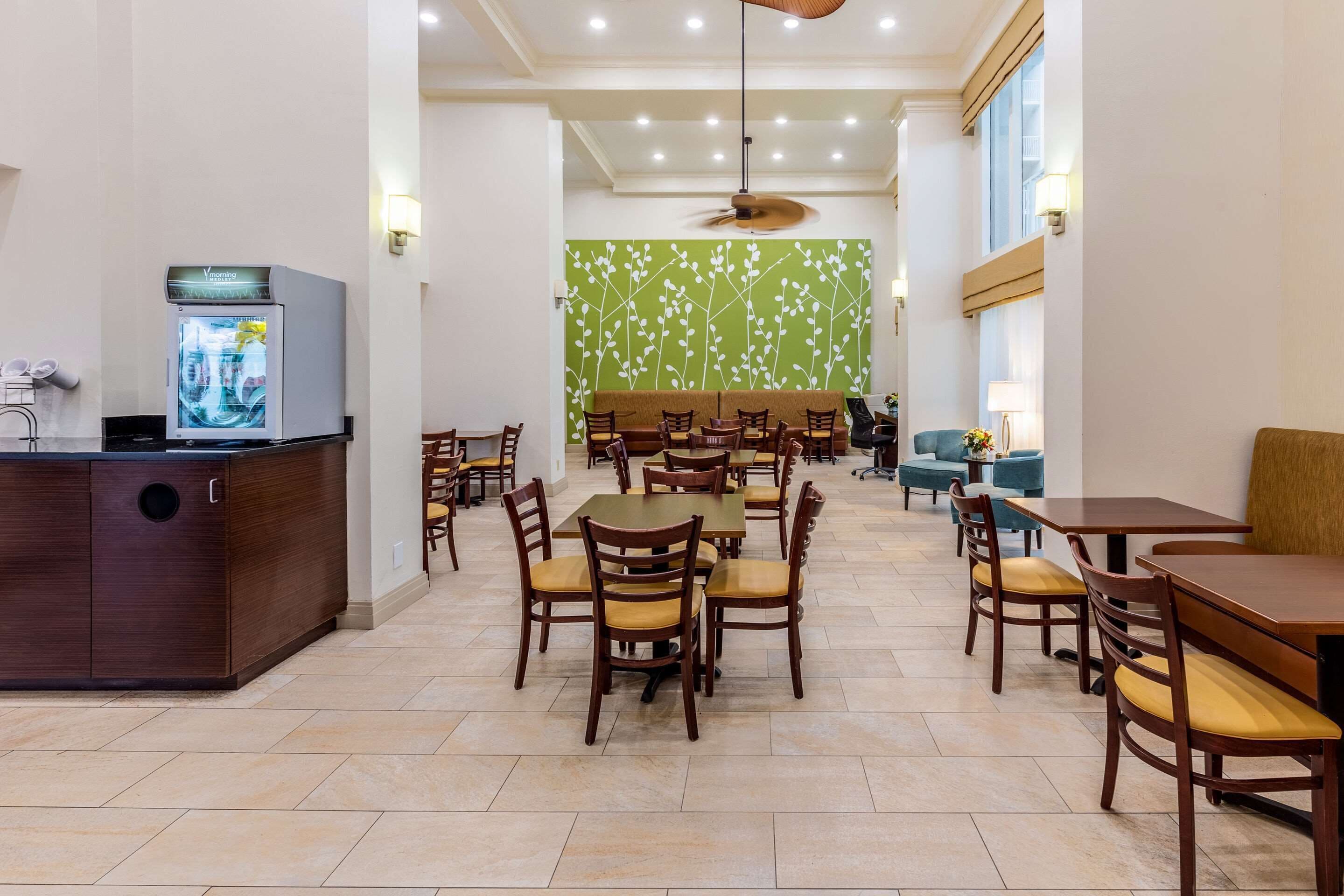 A hotel dining area with wooden tables and chairs, a refrigerator, and a green wall with a floral pattern.