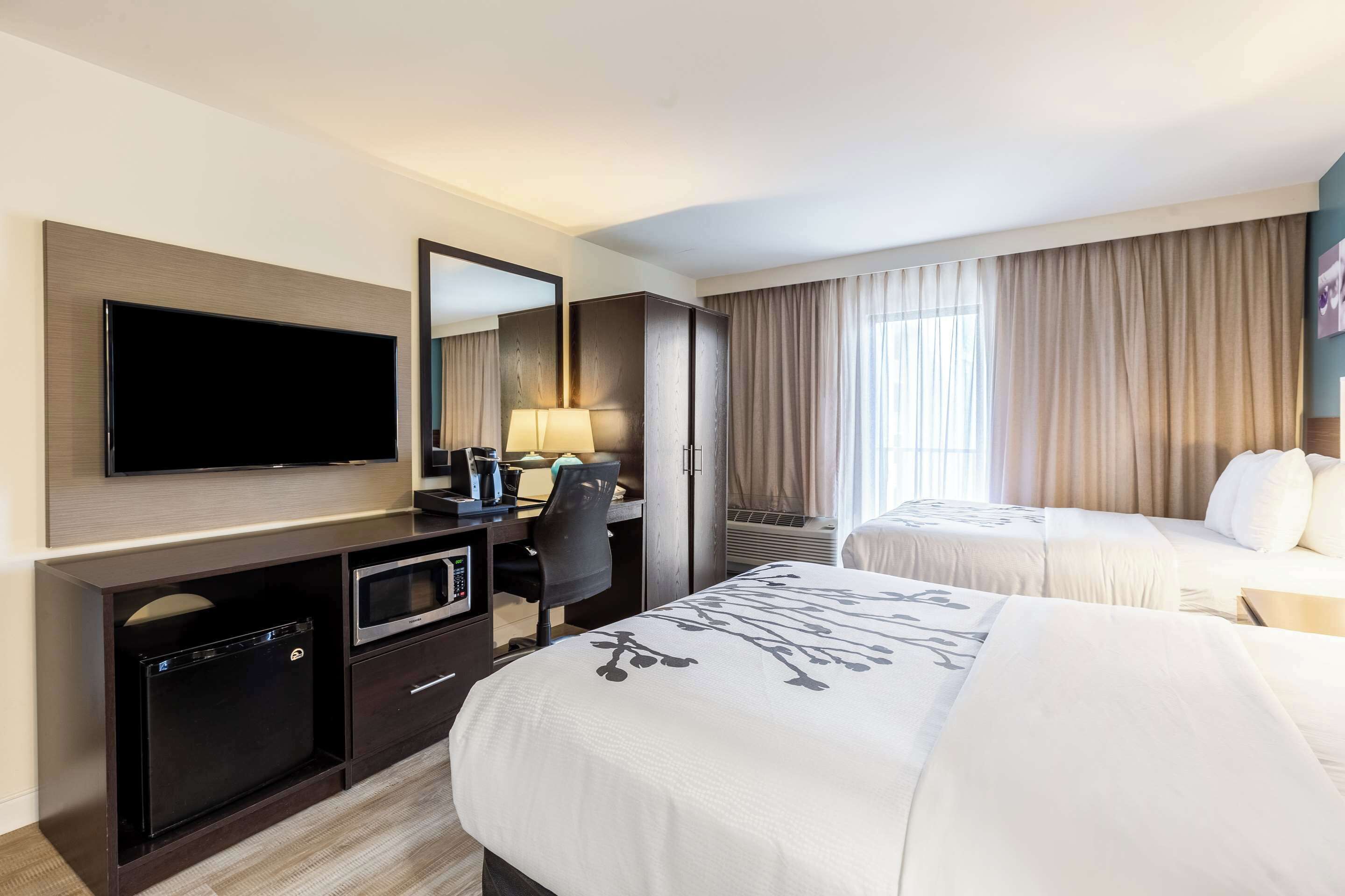 A hotel room with a flat-screen TV, a desk with a lamp, two beds with white bedding, and a large window with curtains.