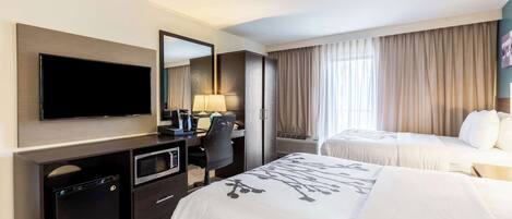 A hotel room with a flat-screen TV, a desk with a lamp, two beds with white bedding, and a large window with curtains.