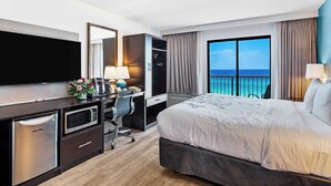 A hotel room with a bed, a desk with a computer, a TV, a minibar, and a large window with a view of the ocean.