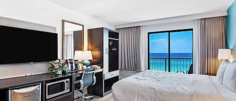 A hotel room with a bed, a desk with a computer, a TV, a minibar, and a large window with a view of the ocean.