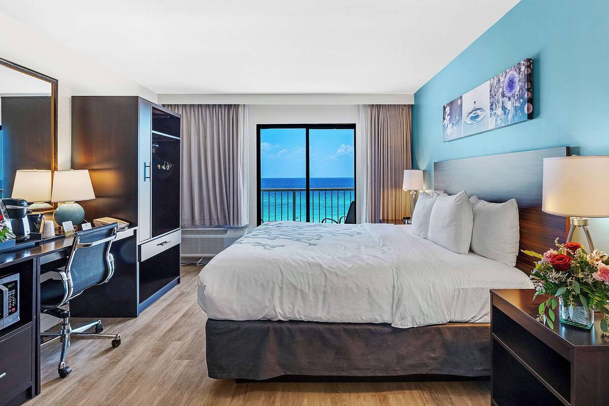 A hotel room with a large bed, a desk with a computer, a TV, and a balcony with a view of the ocean.