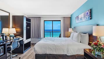 A hotel room with a large bed, a desk with a computer, a TV, and a balcony with a view of the ocean.