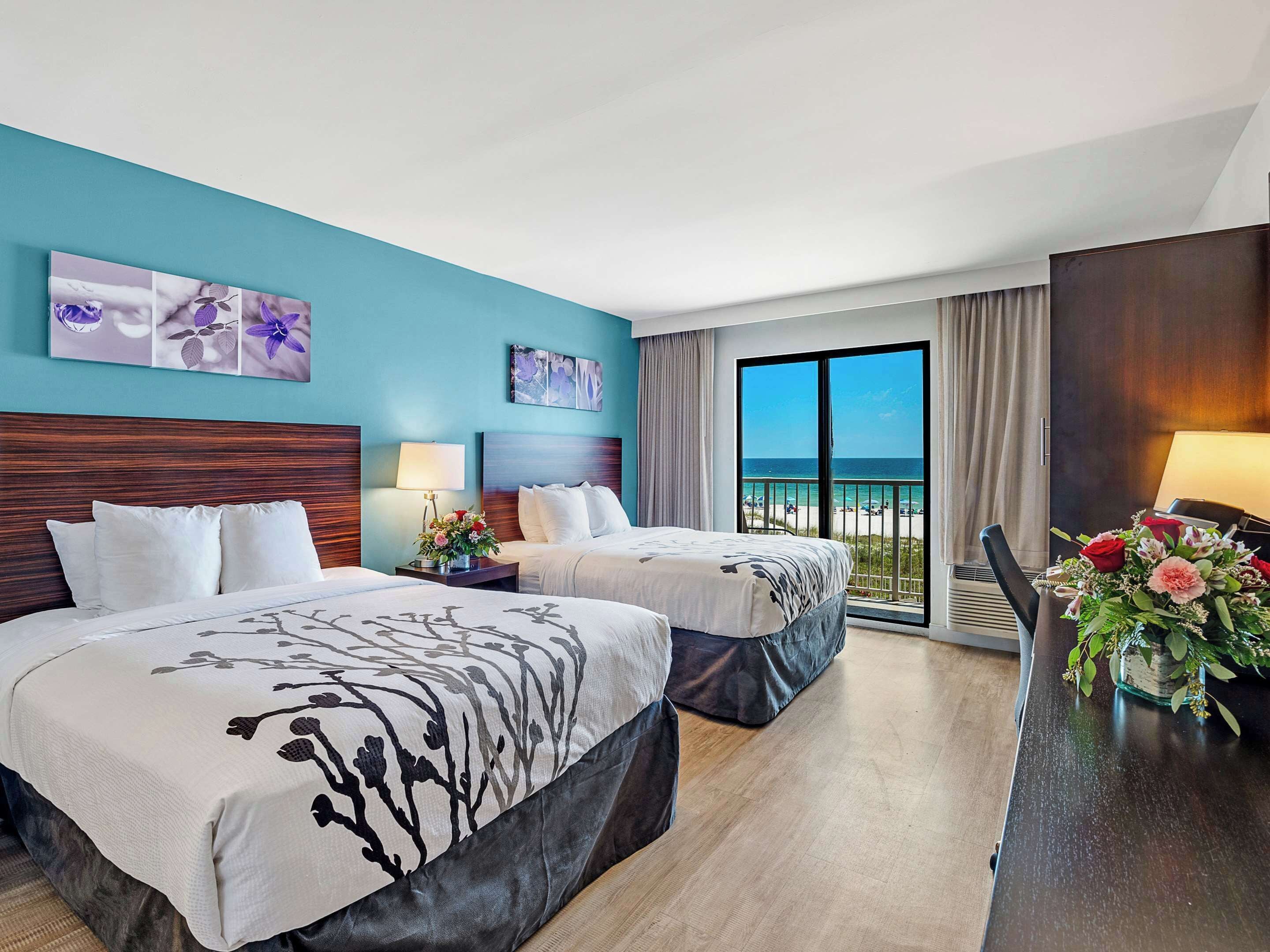 Habitación de hotel con dos camas, un balcón con vista a la playa y obras de arte en las paredes.