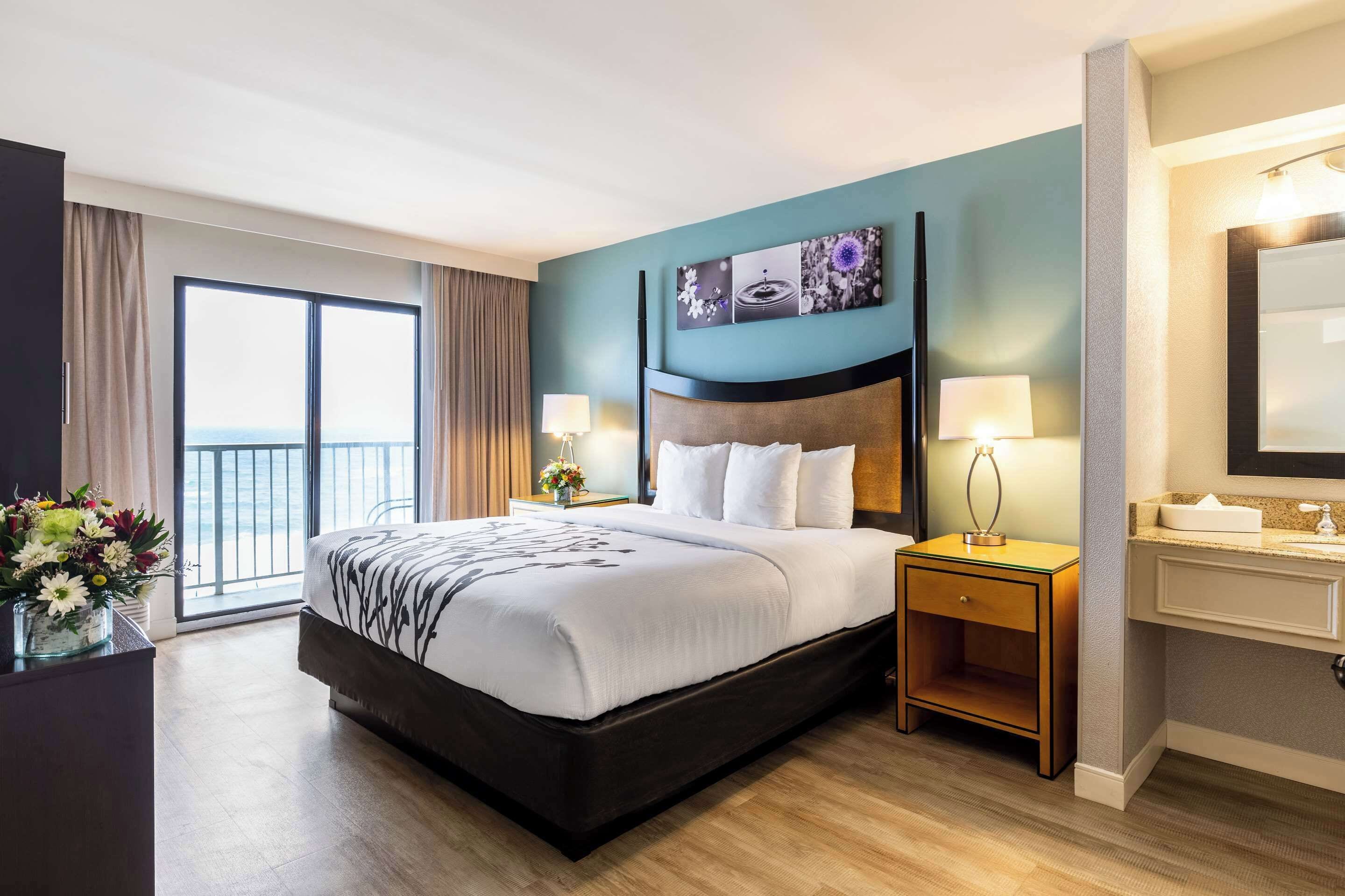 Habitación de hotel con una cama grande, una mesita de noche con lámpara, un baño con espejo y vistas amplias por una ventana grande.