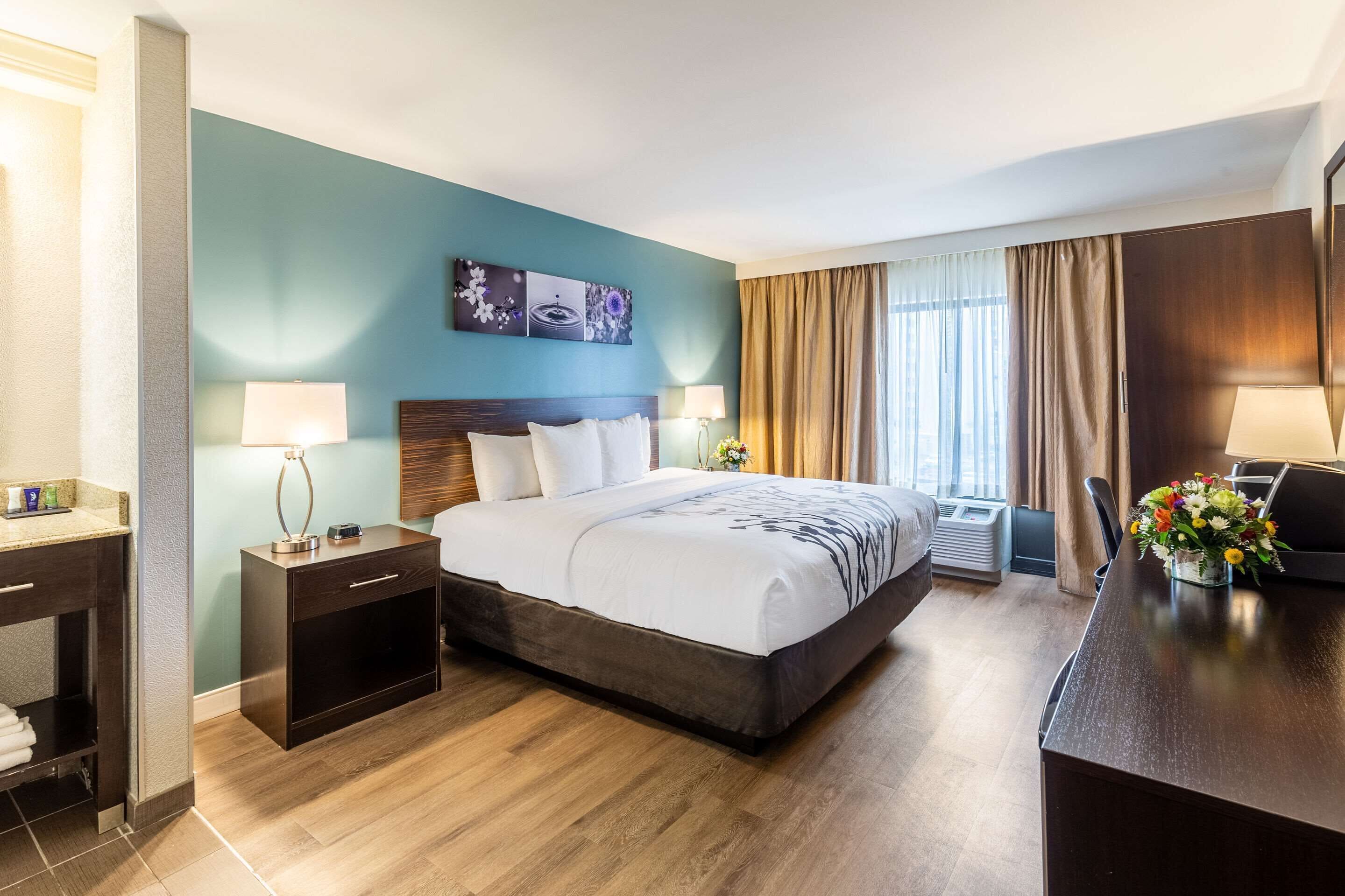 Ett hotellrum med en stor säng, sängbord med lampor, ett skrivbord med en stol, ett litet badrum och ett fönster med gardiner.