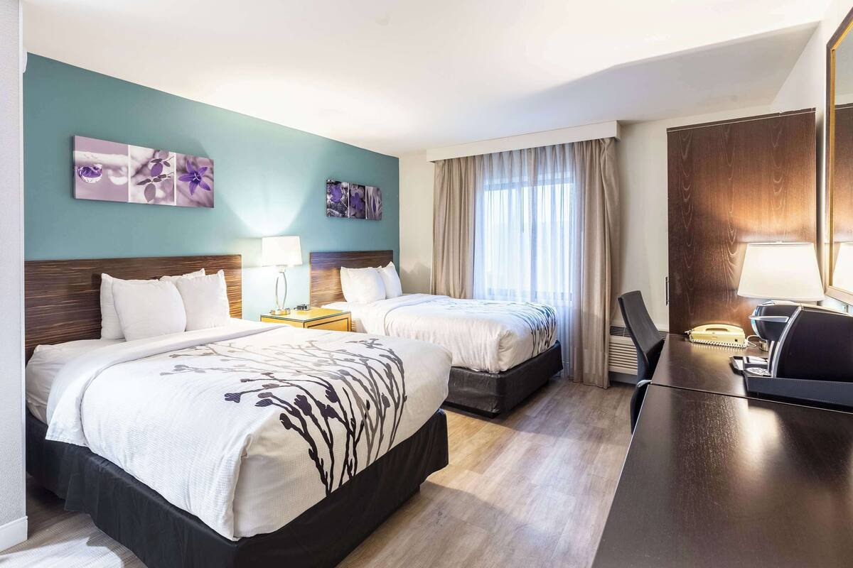 A hotel room with two beds, a desk, and a chair. There are two bedside tables with lamps. The room has a large window with curtains.