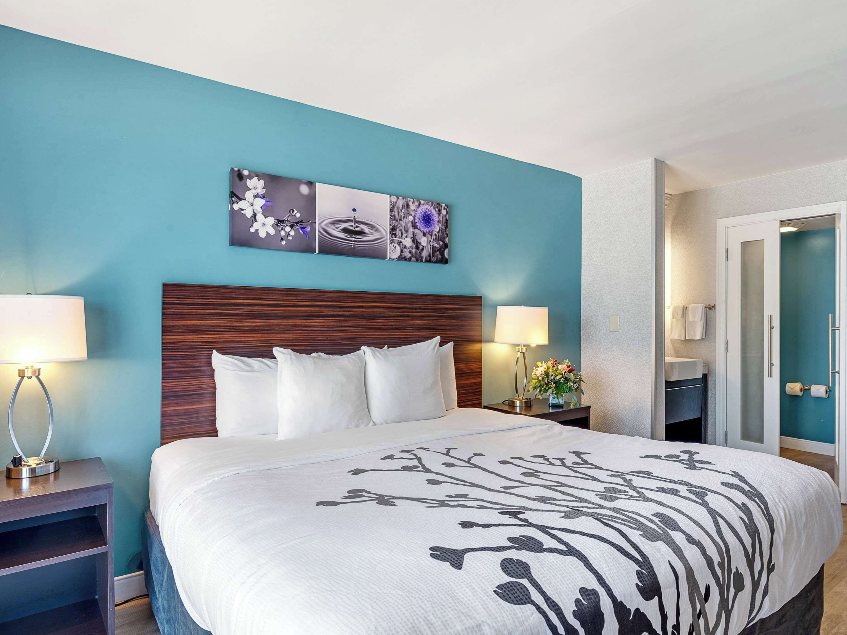 A hotel room with a large bed, two bedside tables with lamps, a nightstand with a vase, and a wall-mounted artwork.