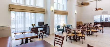 A spacious dining area with multiple tables and chairs, large windows with blinds, and a ceiling fan.