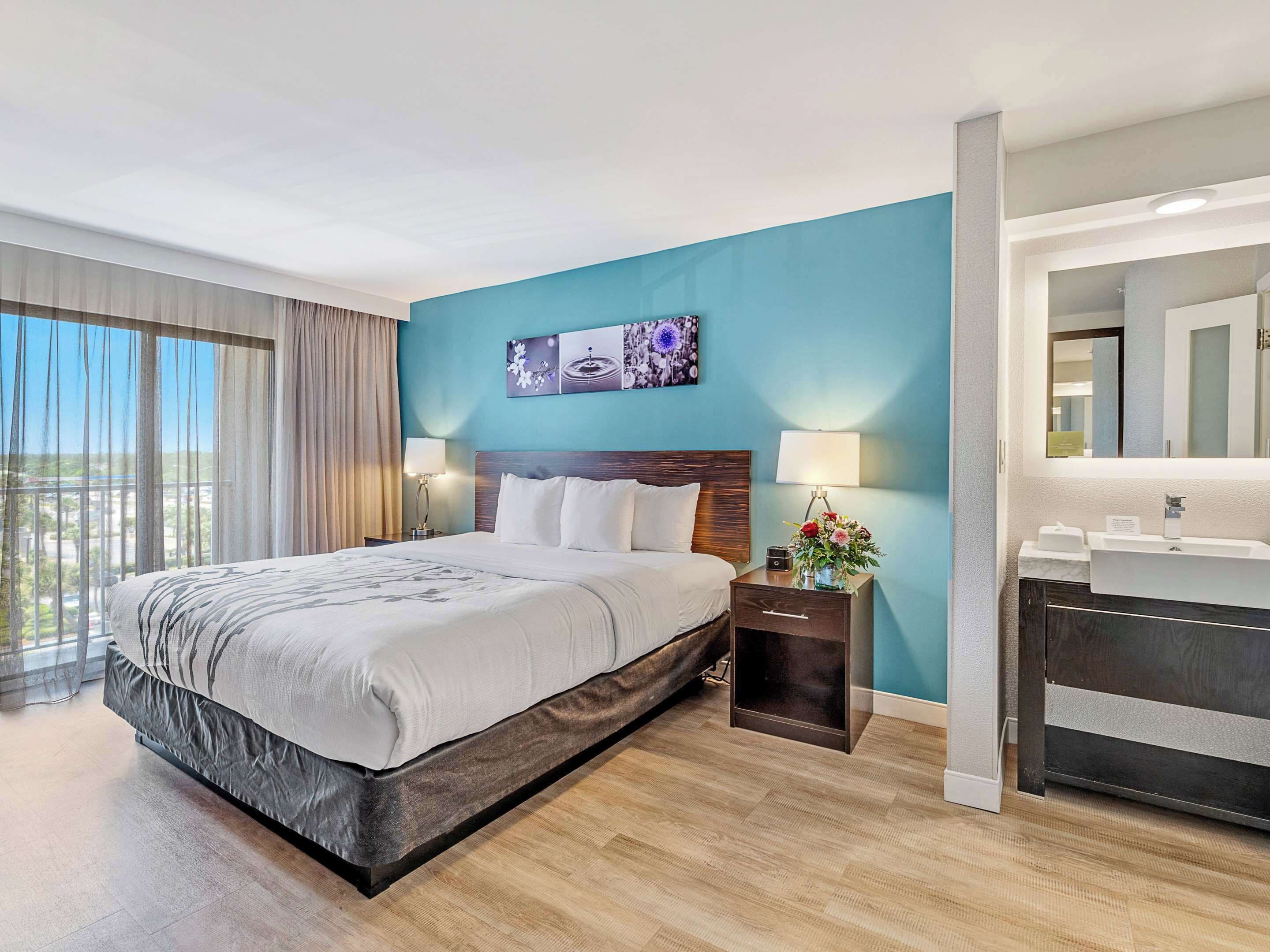A hotel room with a large bed, a nightstand with a lamp, a wall-mounted TV, and a bathroom with a sink and mirror.