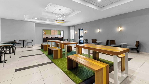 Uma área de refeições moderna com mesas e bancos de madeira, um tapete verde e uma cozinha ao fundo.