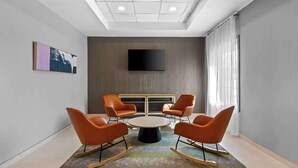 Uma sala de conferências moderna com uma mesa redonda, quatro cadeiras laranja, uma televisão de tela plana na parede e um lustre no teto.