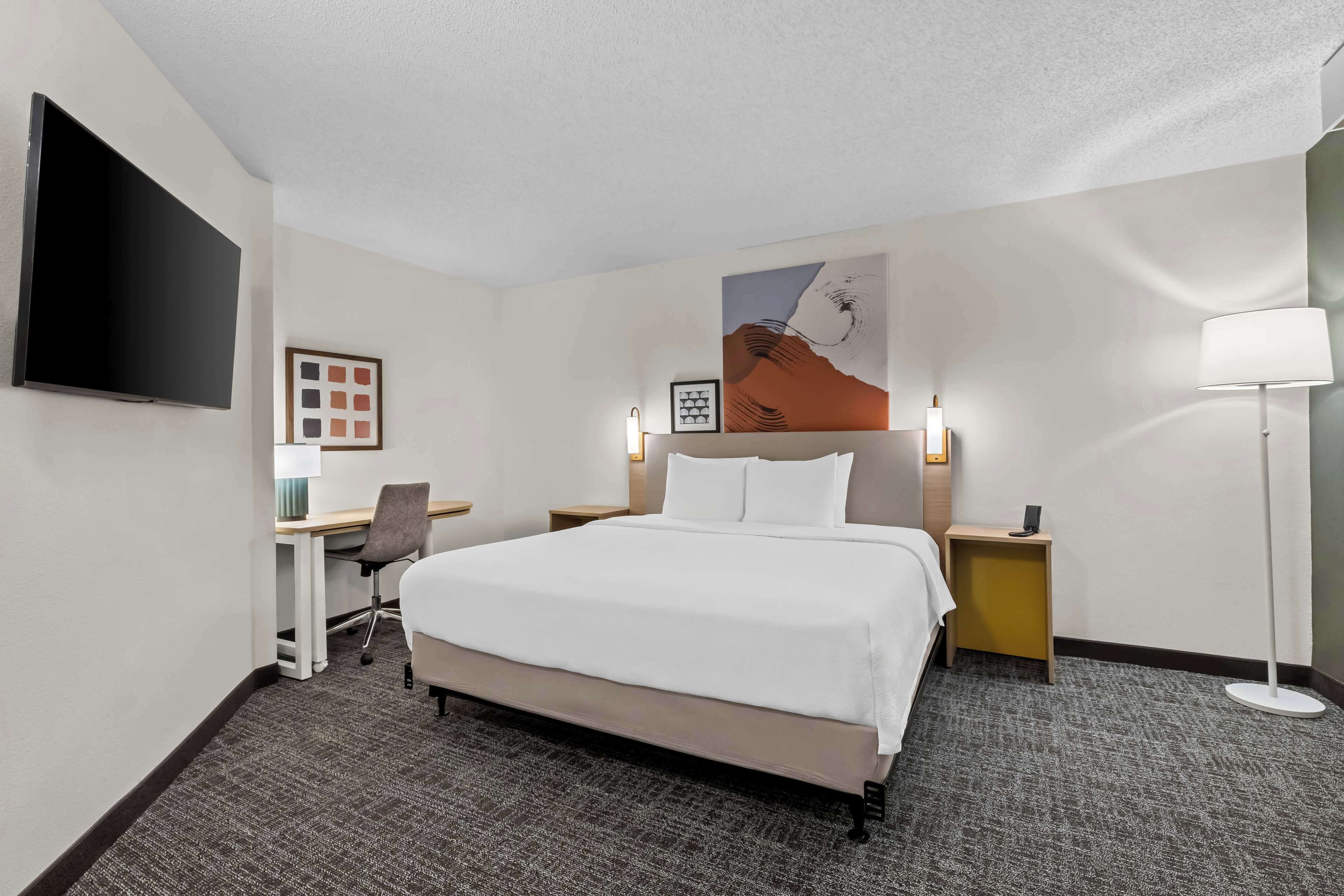 A hotel room with a large bed, a desk, a chair, a flat-screen TV, a painting on the wall, and a floor lamp.