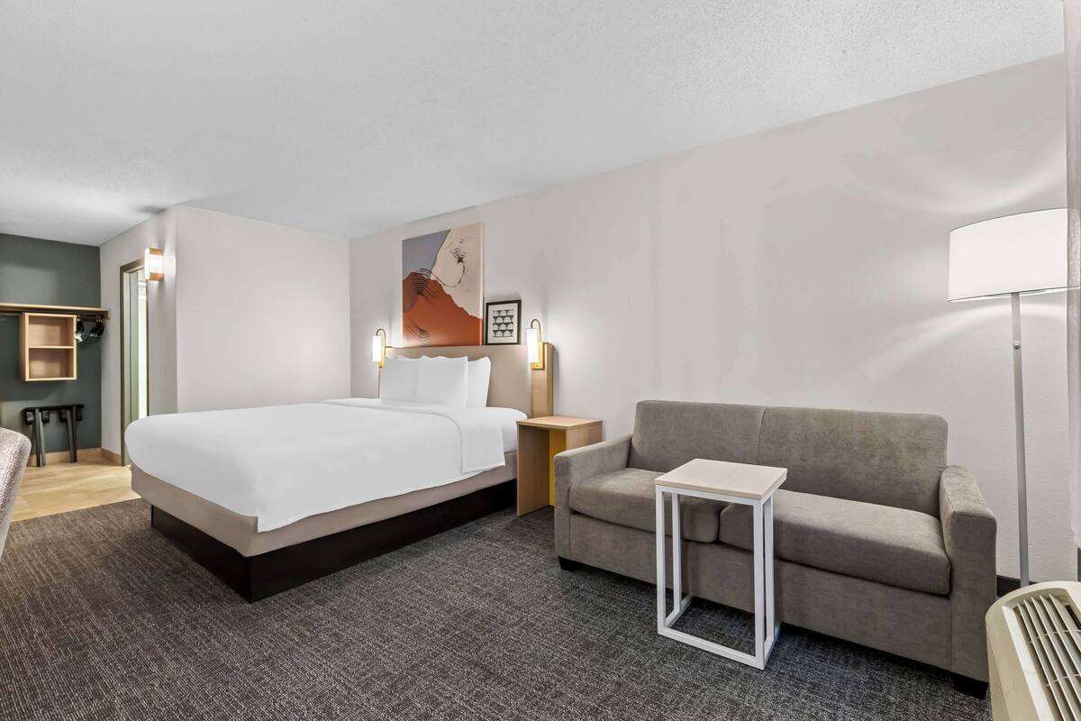 A hotel room with a large bed, a grey sofa, a small table, and a standing lamp.