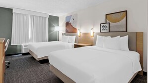 Quarto de hotel com duas camas, uma escrivaninha, uma cadeira, abajur e obras de arte nas paredes.