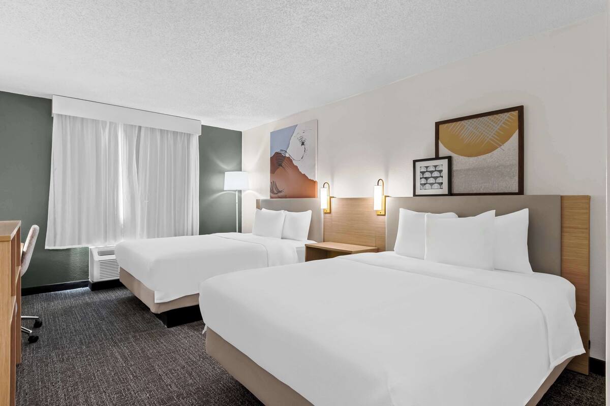 A hotel room with two beds, a desk, a chair, a lamp, and artwork on the walls.
