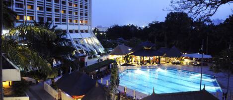 Un hotel con piscina, estructuras de techo de paja y un edificio alto al fondo.