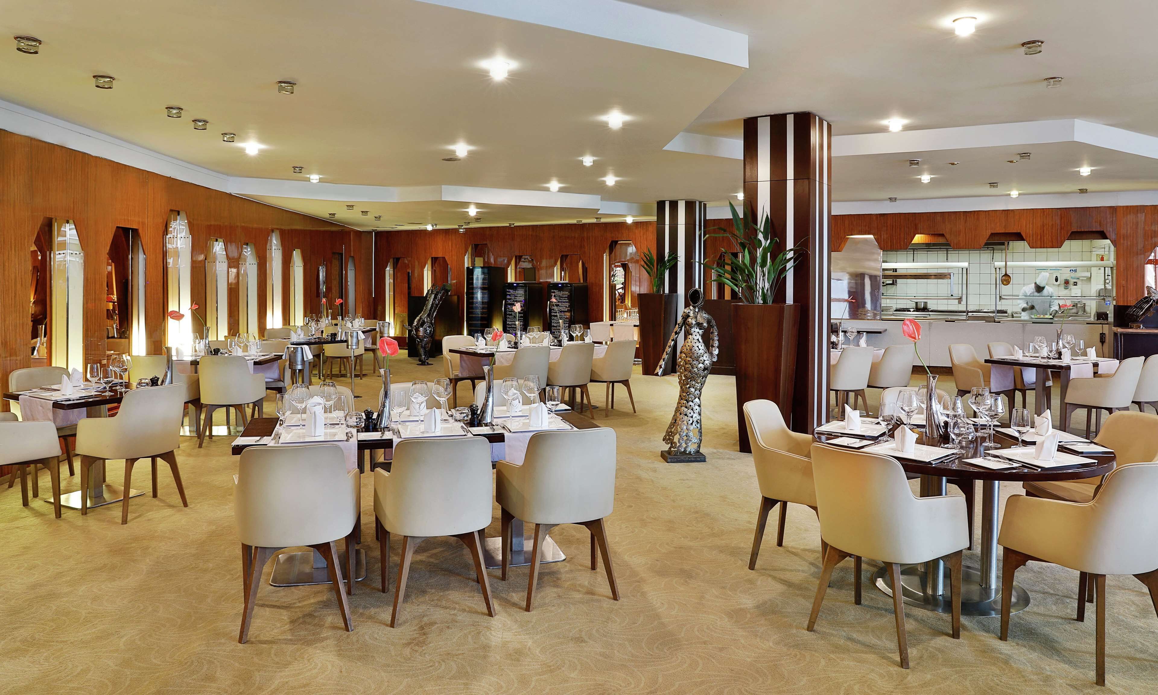 A spacious restaurant with multiple dining tables and chairs, a central column, and a kitchen area visible through a glass partition.