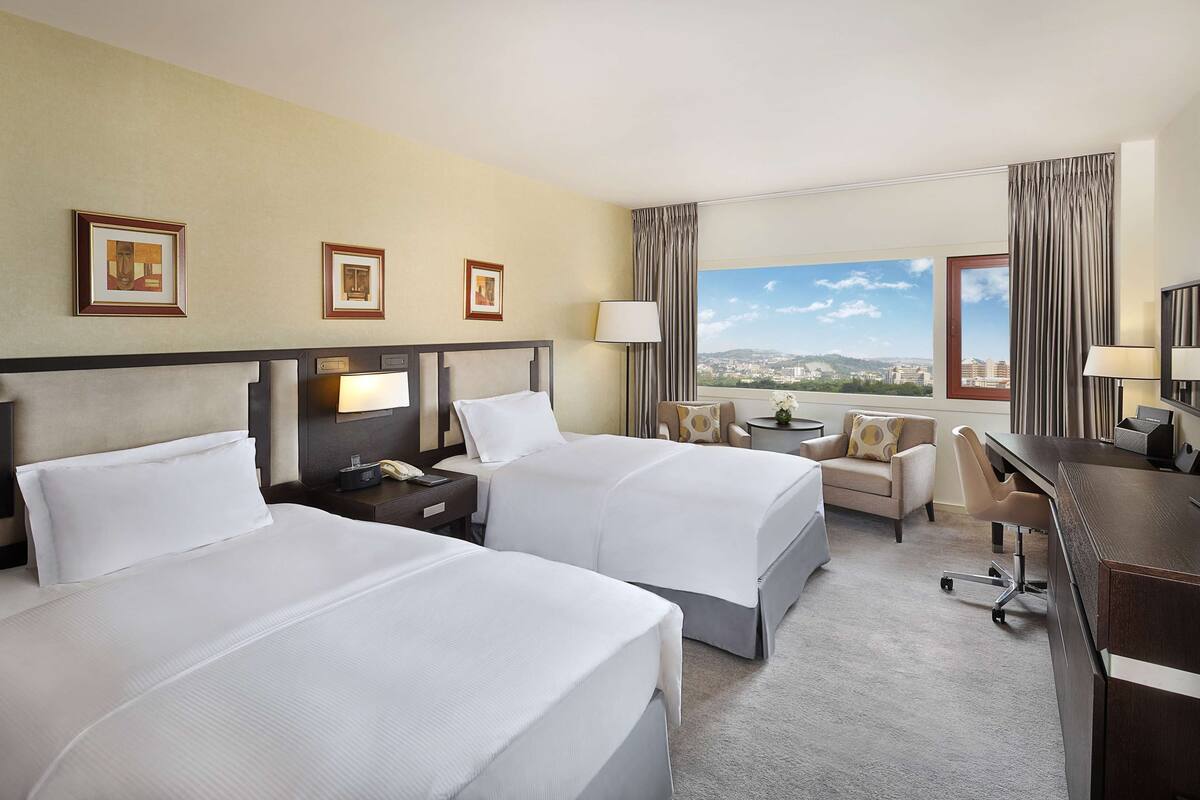 A hotel room with two beds, a desk, a chair, a TV, and a window with a city view.