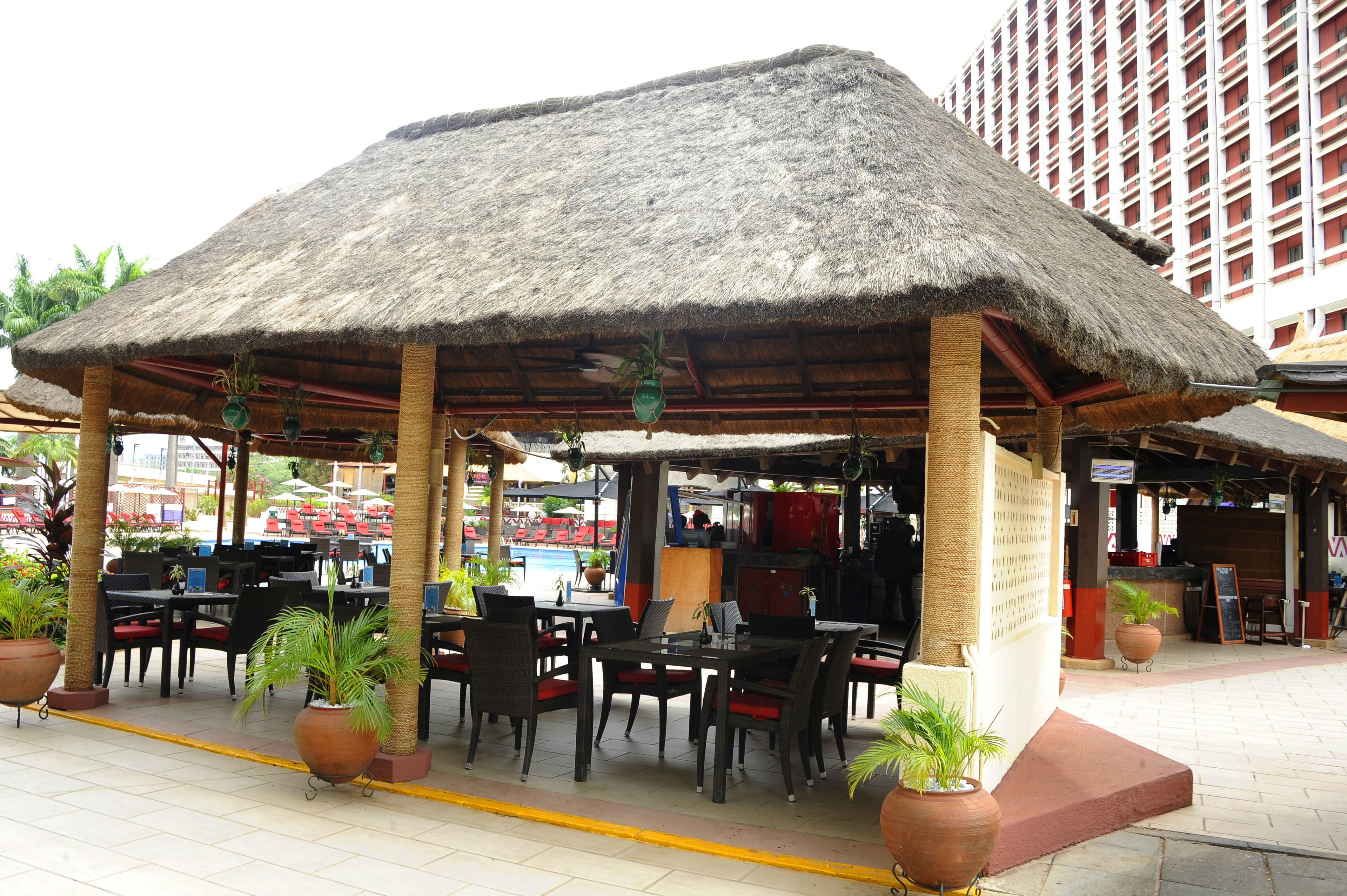 An outdoor seating area with tables and chairs under a thatched roof, surrounded by potted plants.