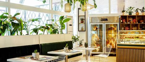 Un café moderno con asientos acolchados verdes, sillas de metal, plantas colgantes y una vitrina de panadería.
