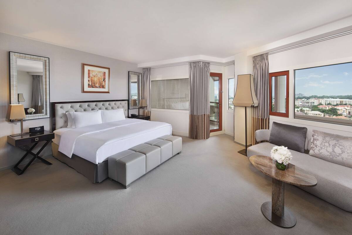 A hotel room with a large bed, a sofa, a small table, a mirror, and a view of the city.
