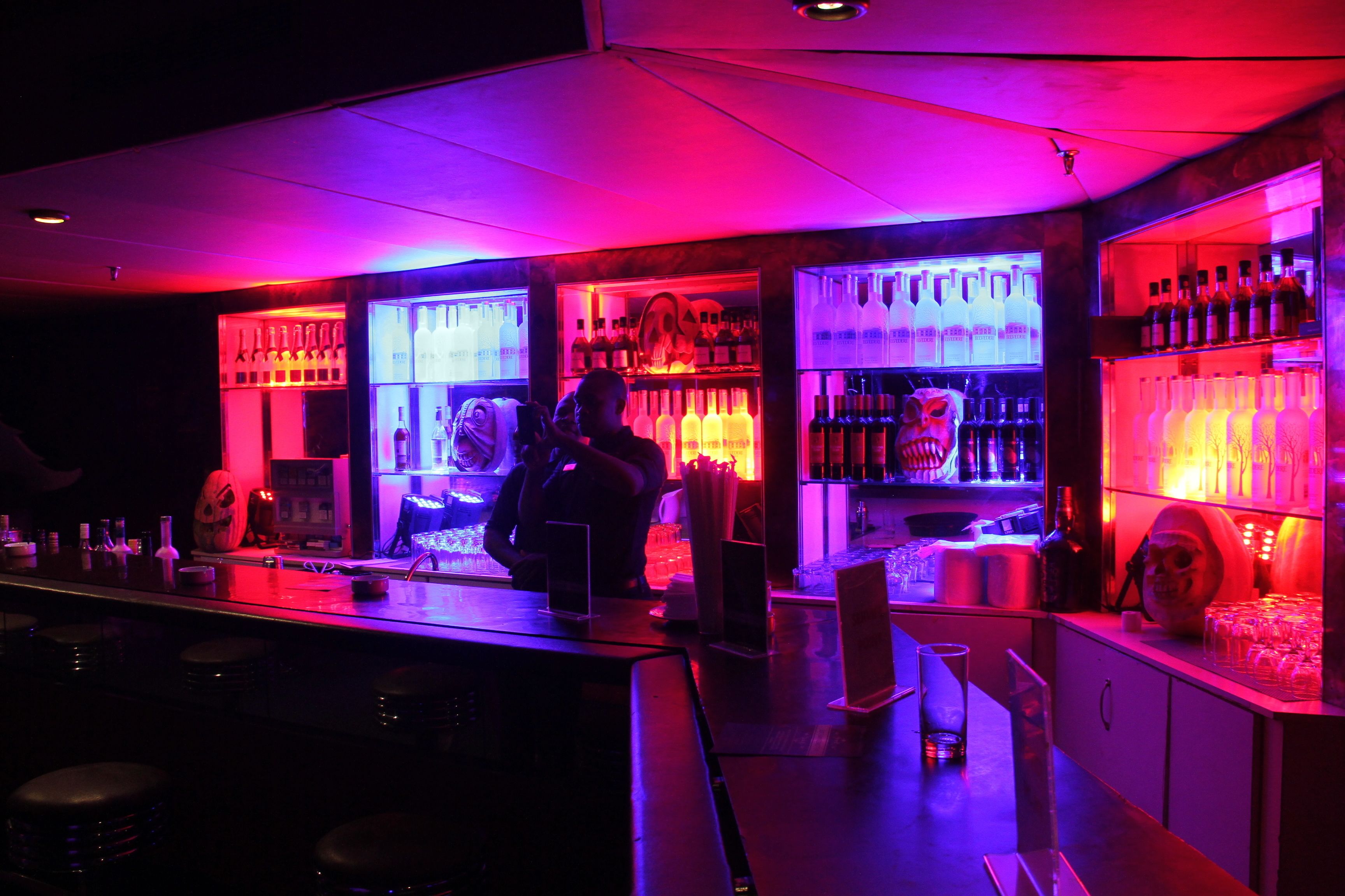 A bar area with illuminated shelves of bottles and glasses, a bar counter, and stools.
