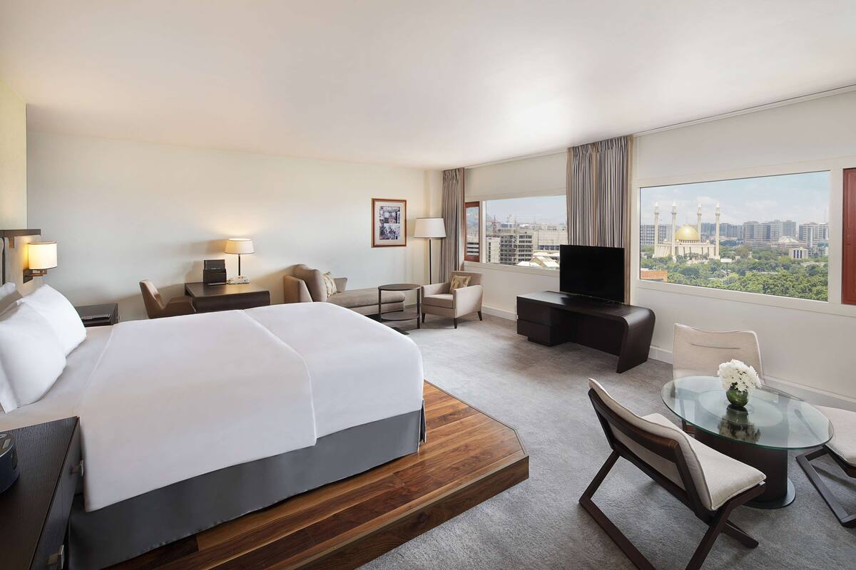 A hotel room with a large bed, a TV, a desk with a lamp, a chair, a small table with a vase, and a view of the city.