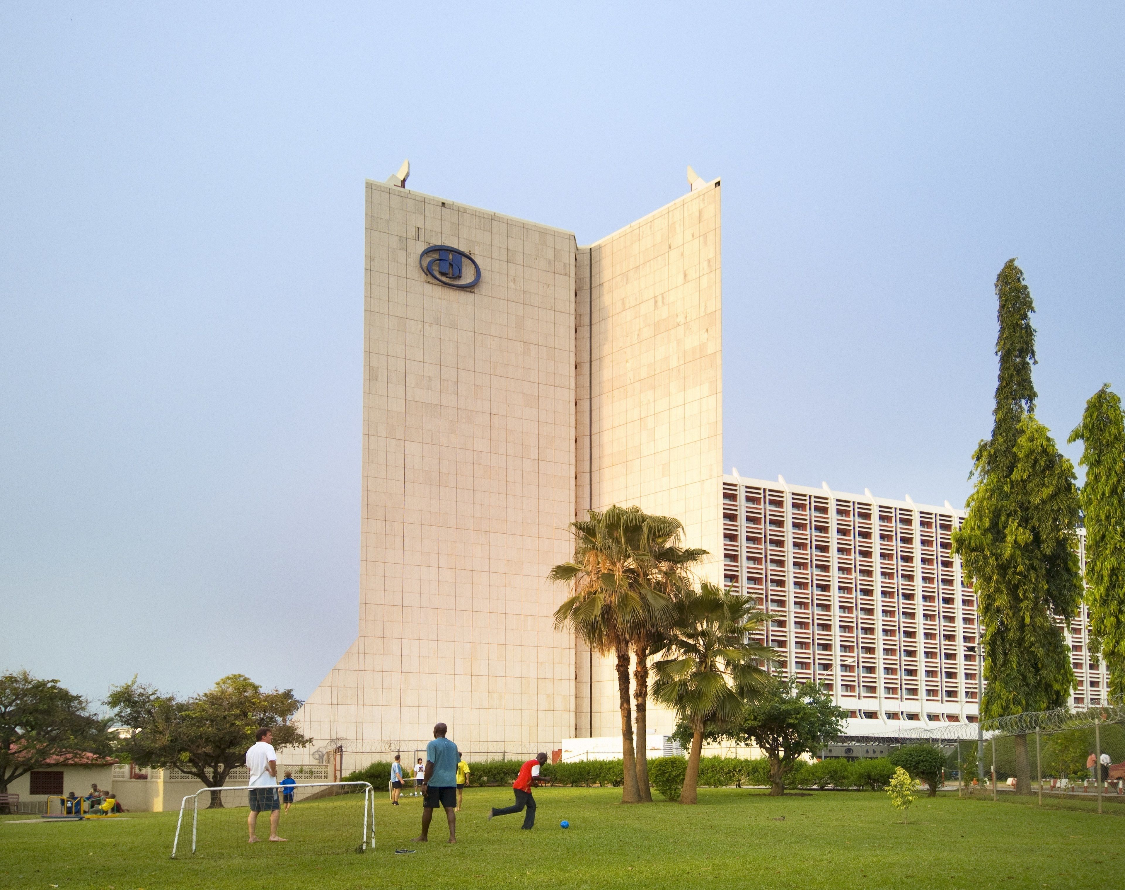A tall building with a distinctive logo, surrounded by greenery and palm trees.