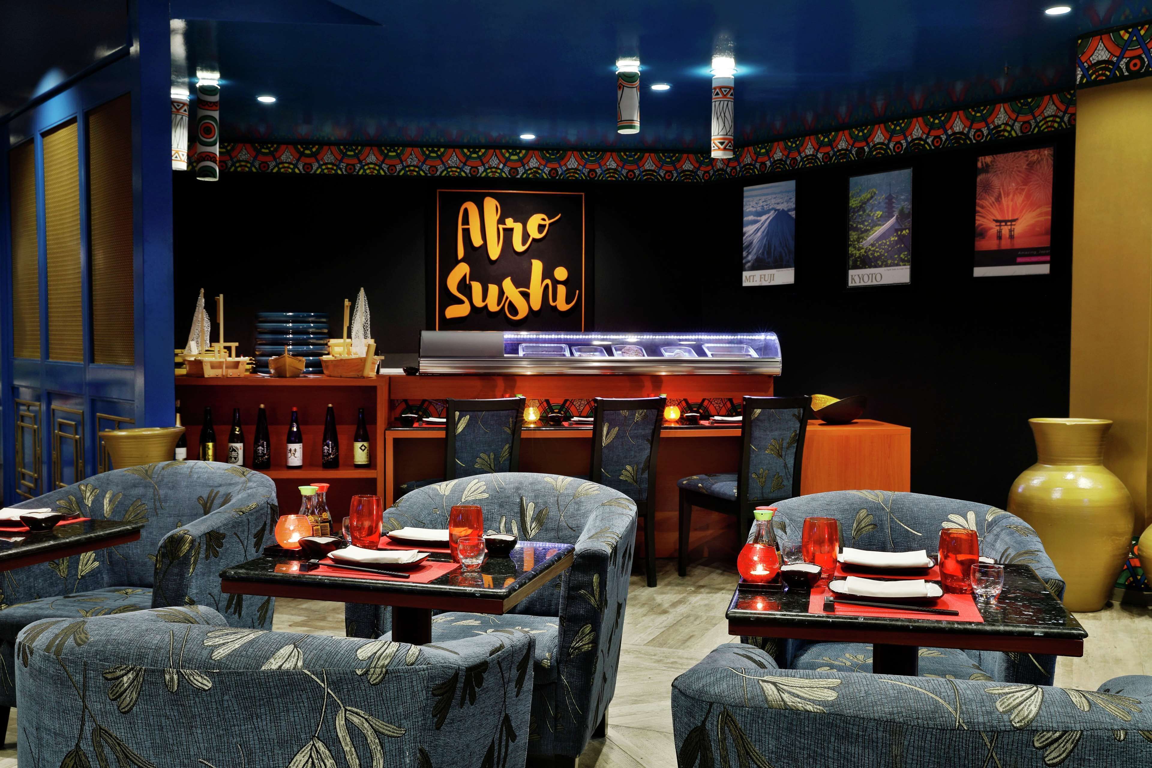 Ein Restaurantinneres mit blau gepolsterten Stühlen, Tischen, die mit roten Servietten und Gläsern gedeckt sind, und einem Neon-Schild mit der Aufschrift 'Alro Sushi'.