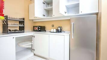 A compact kitchen with white cabinets, a stainless steel refrigerator, a coffee maker, and a basket under the counter.