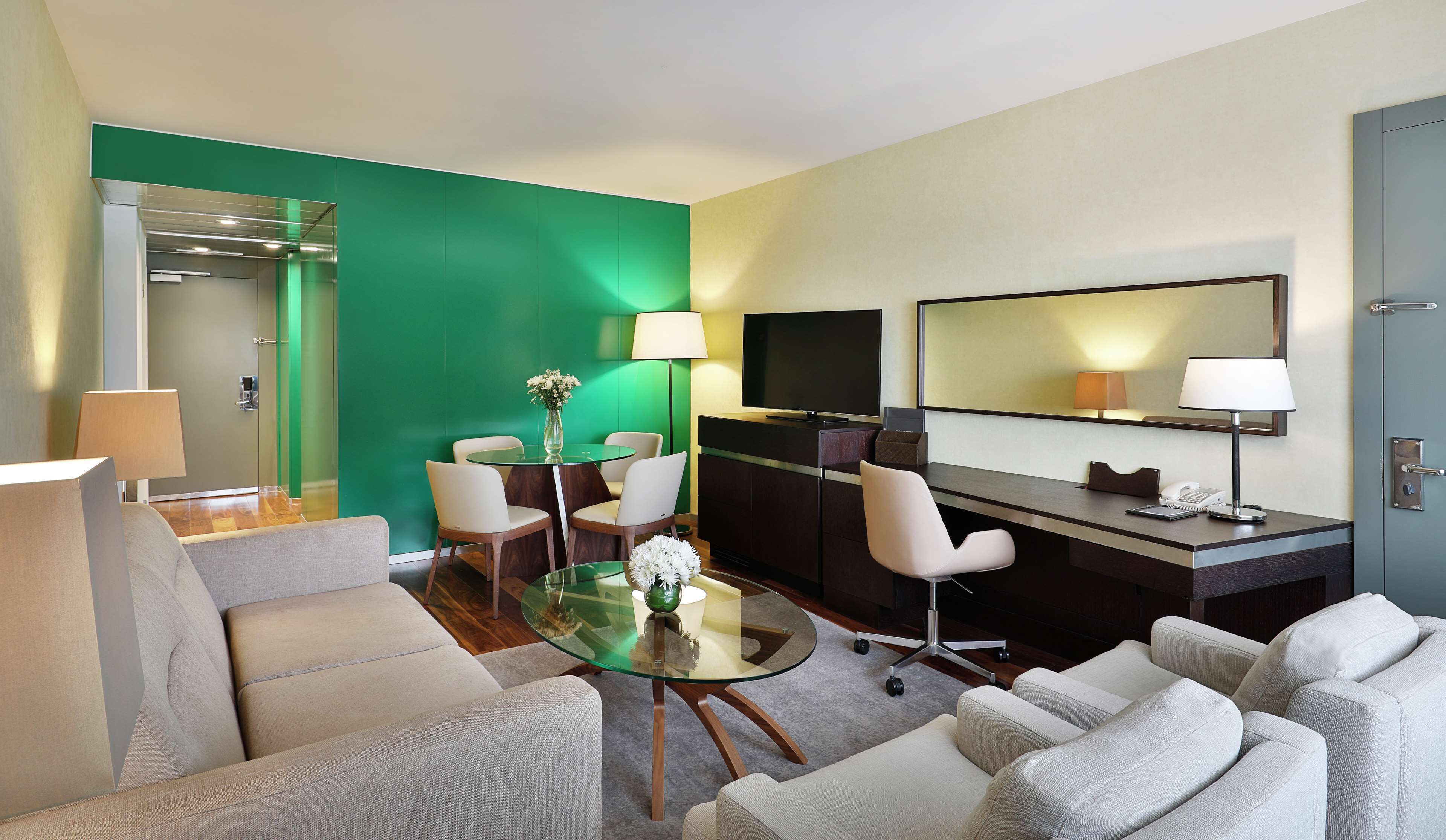 A modern hotel room with a green accent wall, a glass dining table, a sofa, a desk with a TV, and a mirror.