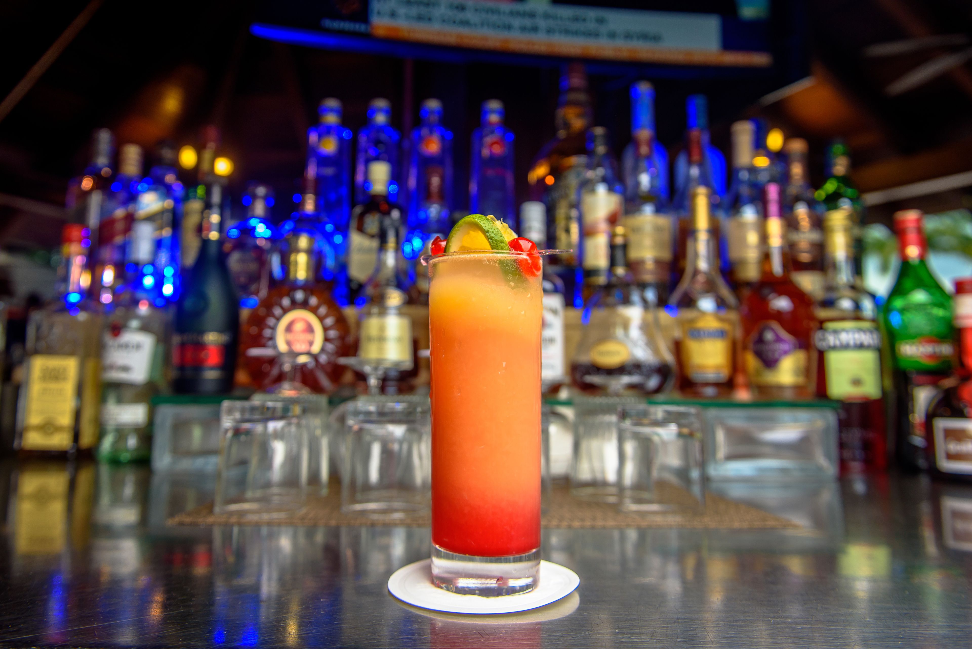 A bar setting with a tall glass of a red drink garnished with a lime and a cherry, surrounded by various bottles of alcohol.