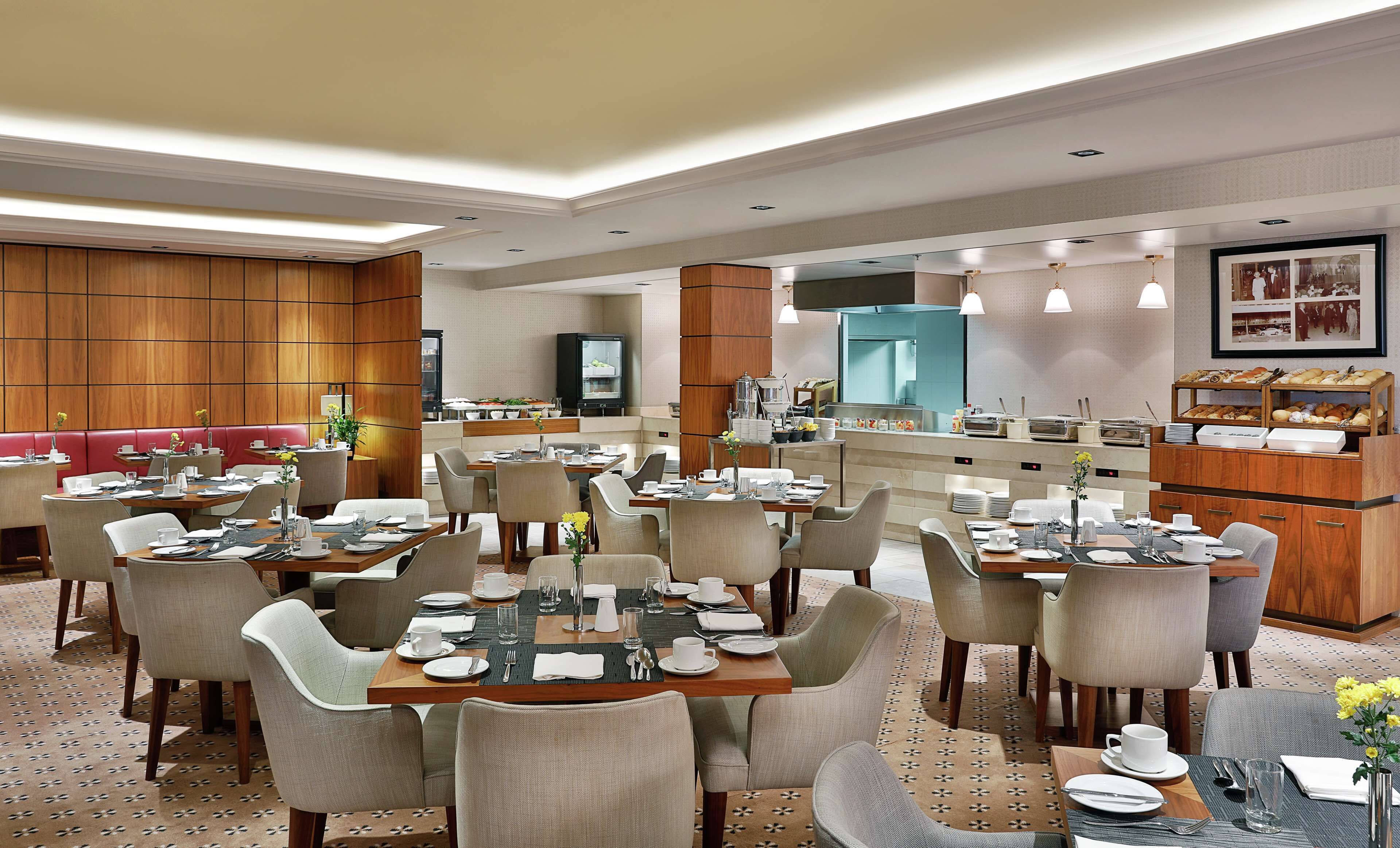 A hotel dining area with multiple tables set for breakfast, a kitchen in the background, and a buffet counter with various food items.