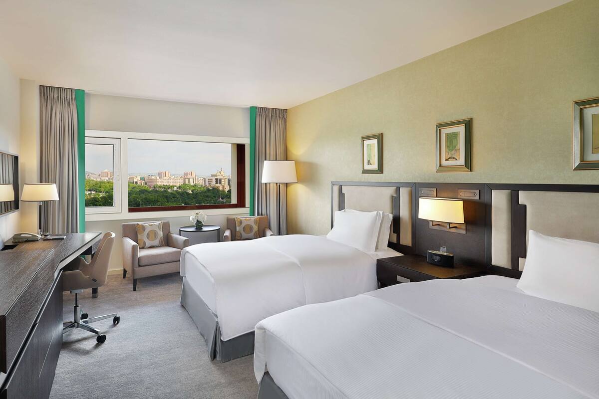 A hotel room with two beds, a desk, a chair, a TV, and a view of the city.