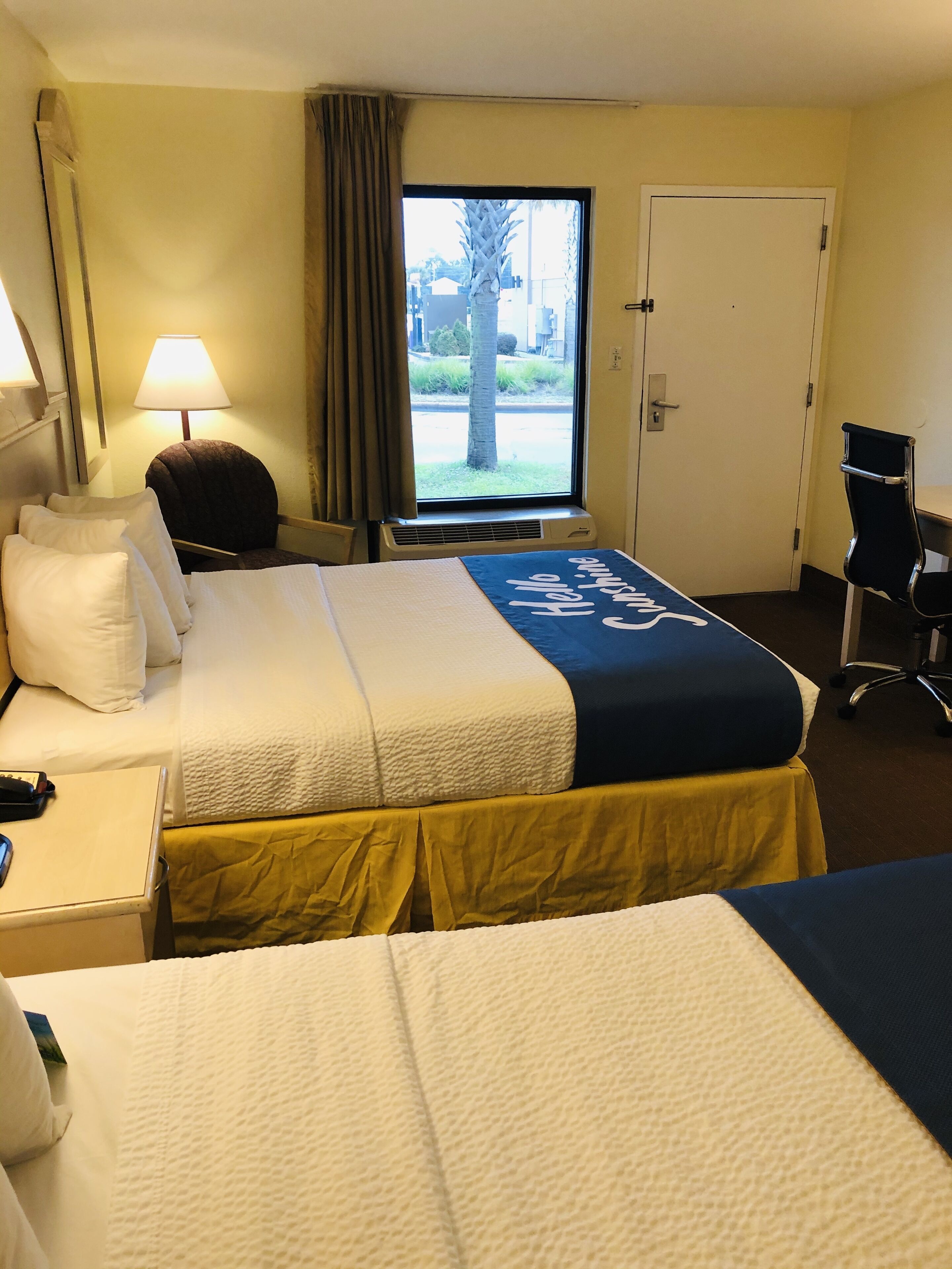 Une chambre d’hôtel avec deux lits, un bureau, une chaise, une lampe et une fenêtre donnant sur l’extérieur.