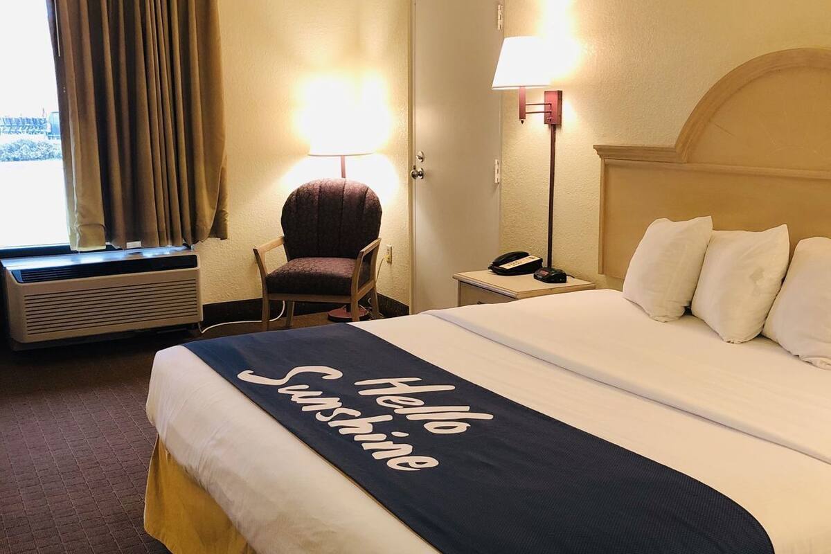Une chambre d’hôtel avec un lit, une chaise, un bureau avec une lampe, un téléphone et une fenêtre avec des rideaux.